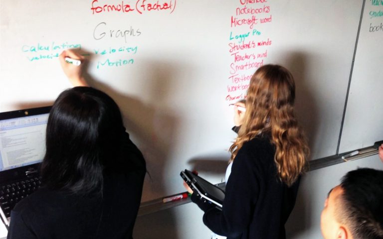 2 female students holding computers writing formulas on whiteboard