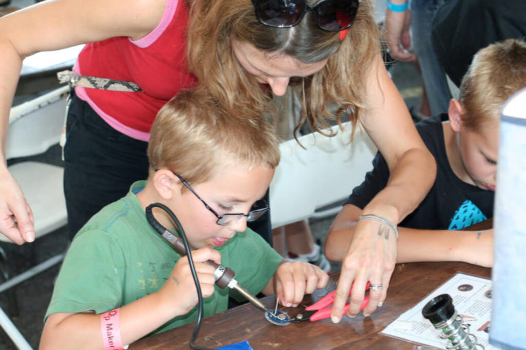 Woman standing over the shoulder of a child while he solders something