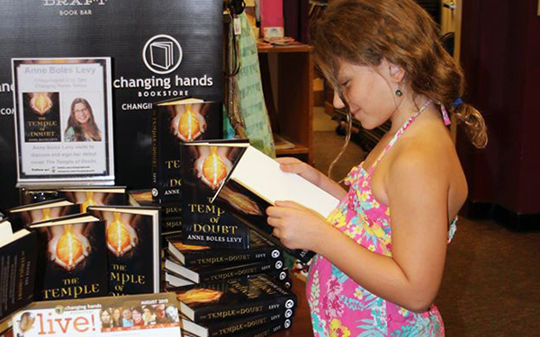 young girl reading book while standing in front of other books