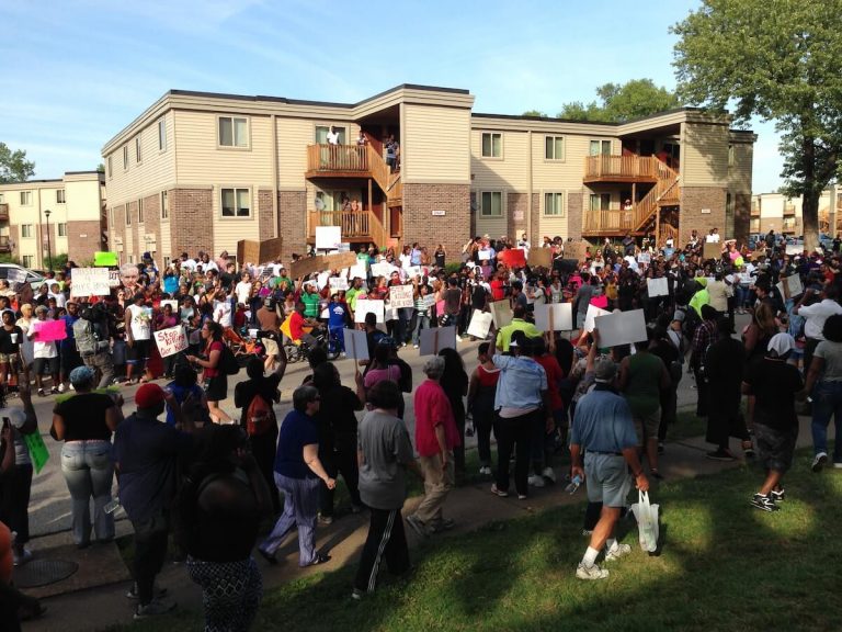 A large crowd with signs protests outside. A tan building is in the background.