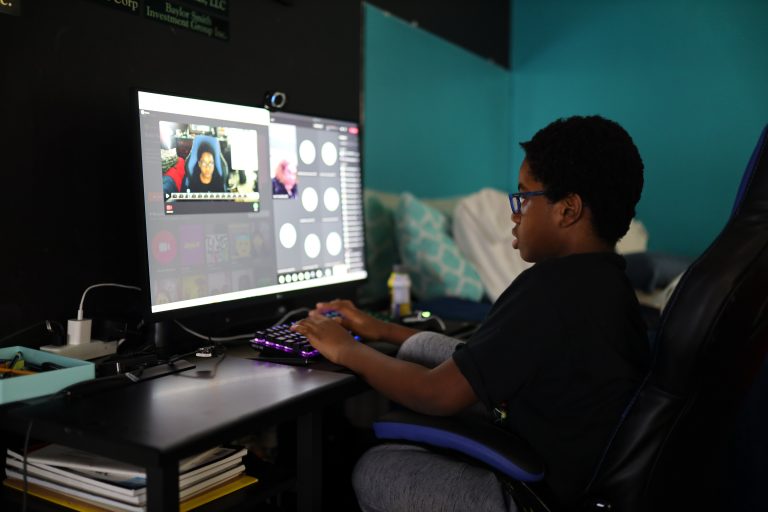 A young Black boy with glasses types and sits in front of a large computer monitor.