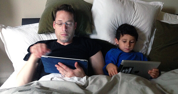 Dad and son in bed both looking at tablets